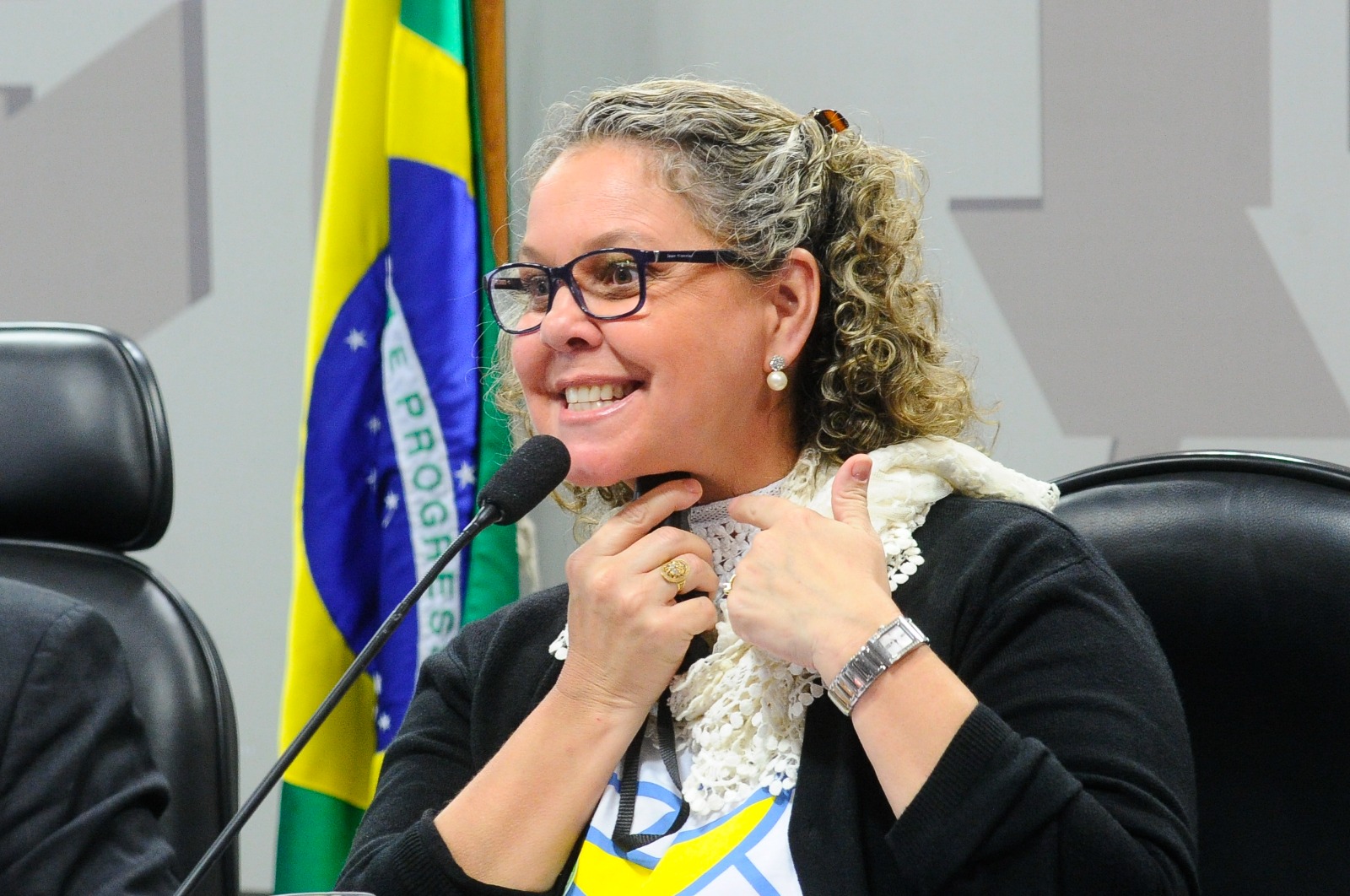 Melissa Medeiros sorrindo durante audiência pública, com a bandeira do Brasil ao fundo